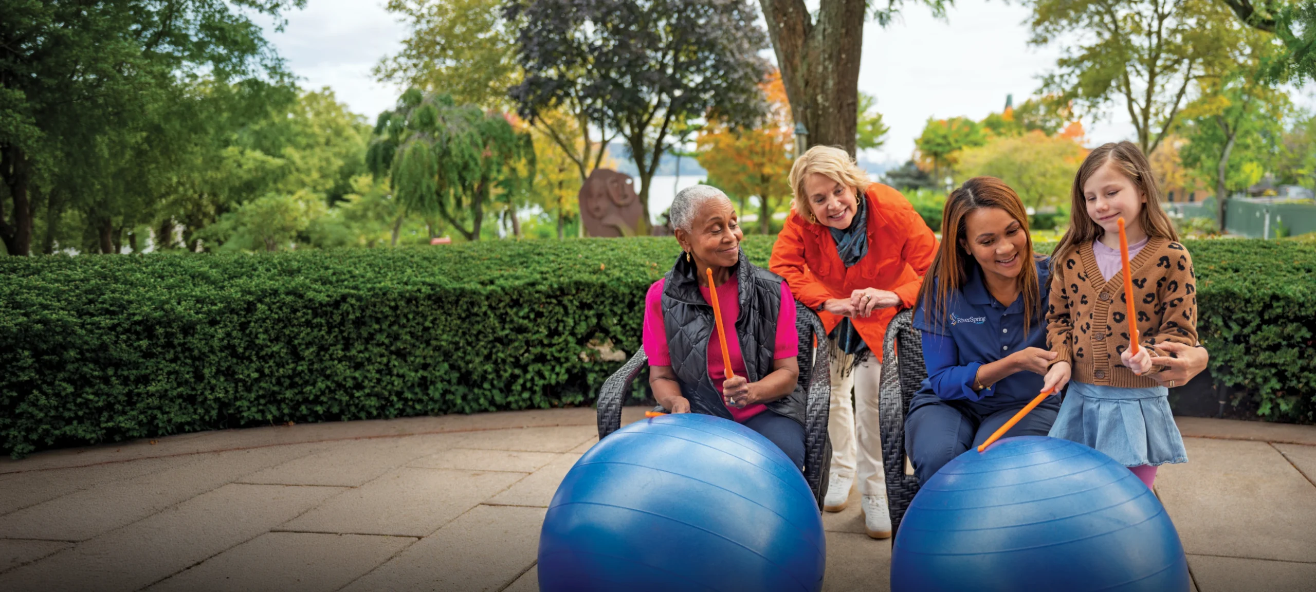 Two women, a RiverSpring Living caregiver, and a young girl sit outdoors with exercise balls, smiling together.
