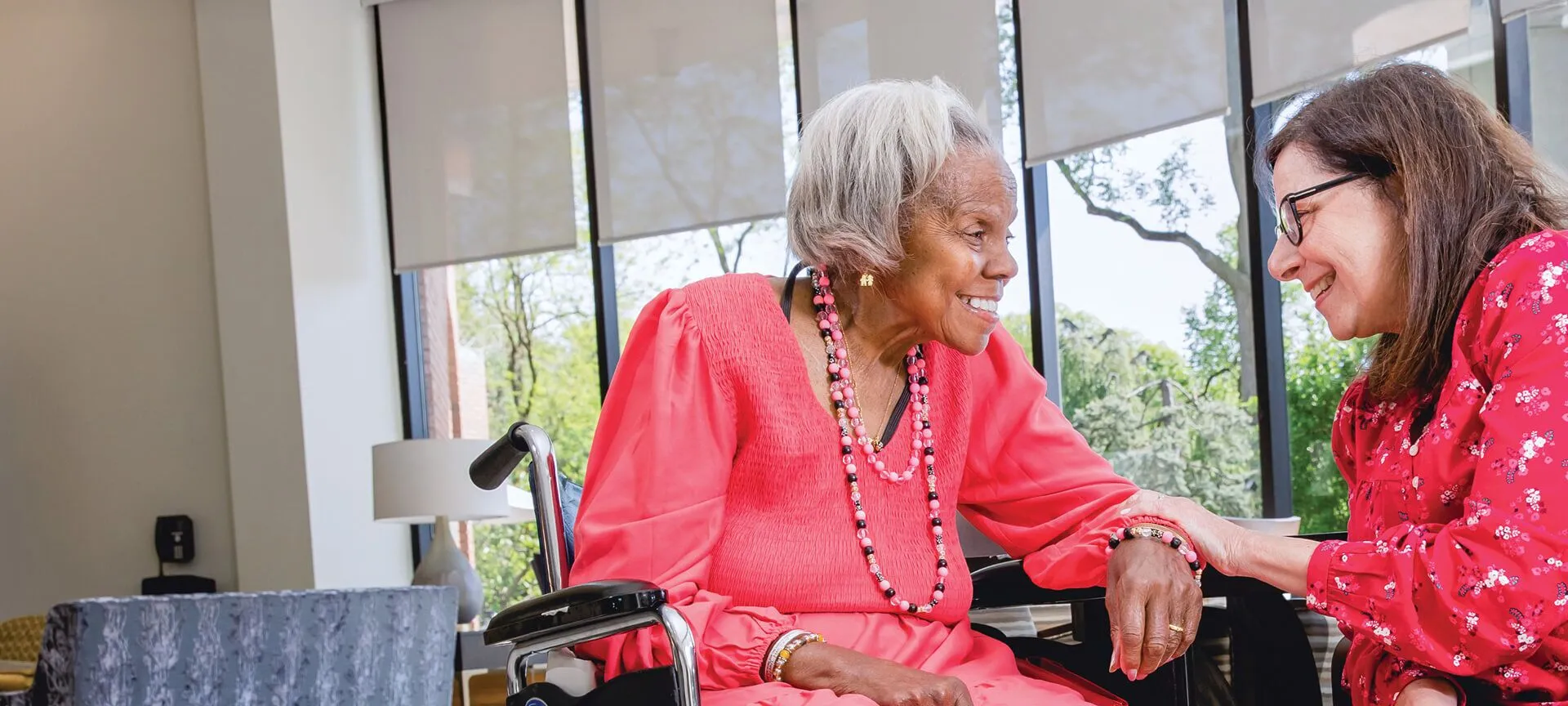 A Certified Home Health Aide and a woman sit together indoors, smiling and holding hands by large windows with trees visible outside.