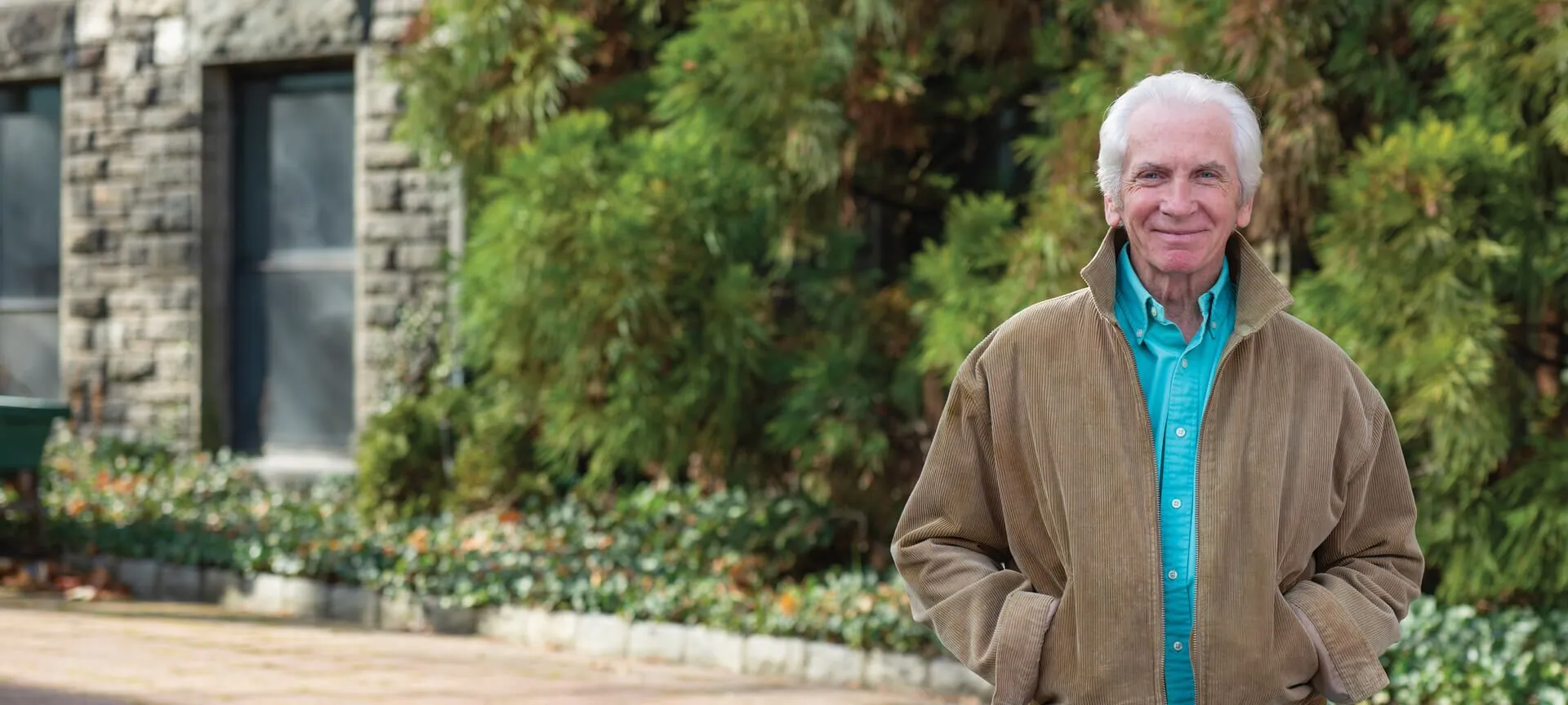 An older man with white hair stands outside in front of a stone building and green bushes, wearing a tan jacket over a teal shirt with his hands in his pockets—embracing the freedom of Independent Living.