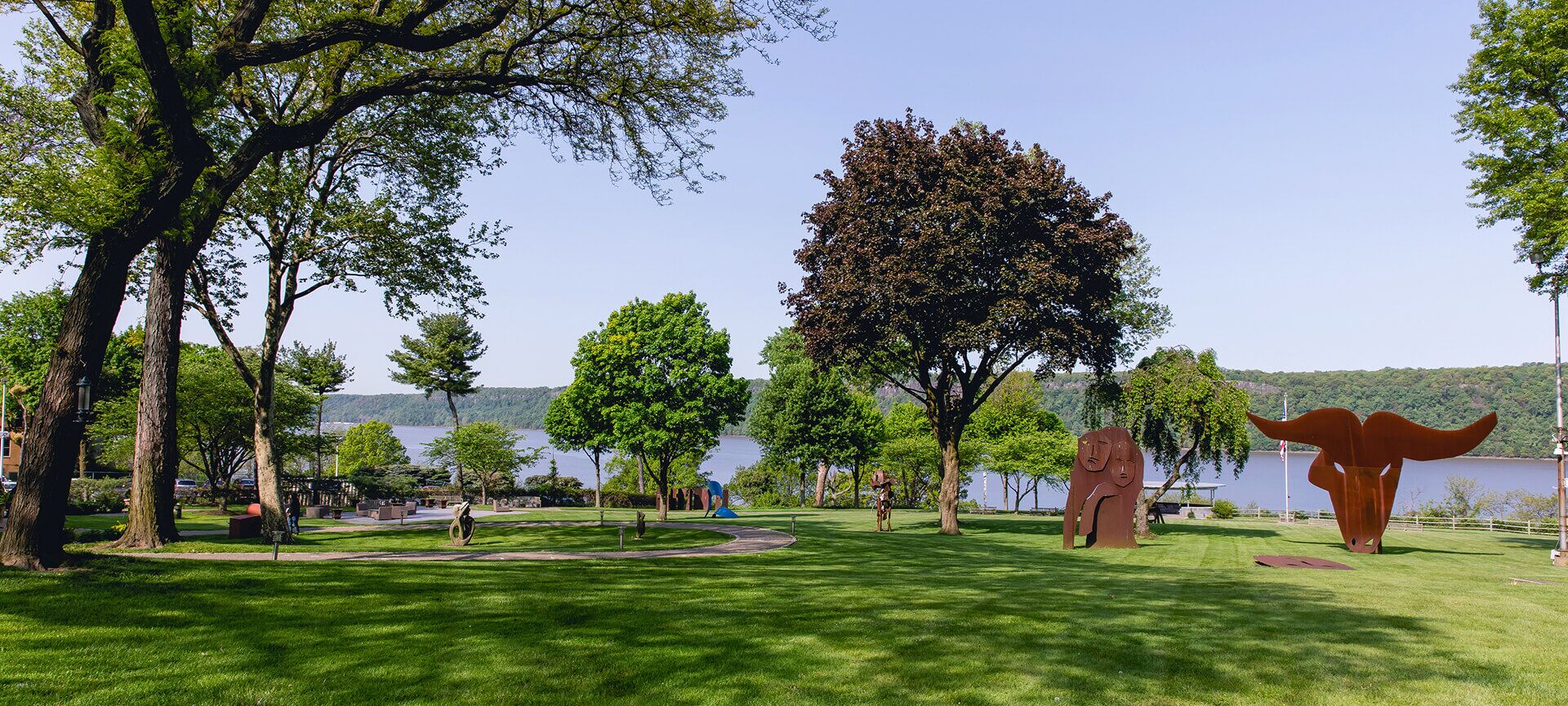 A grassy park with large trees, modern metal sculptures, and a view of a river and distant wooded hills under a clear sky, perfect for outdoor relaxation.