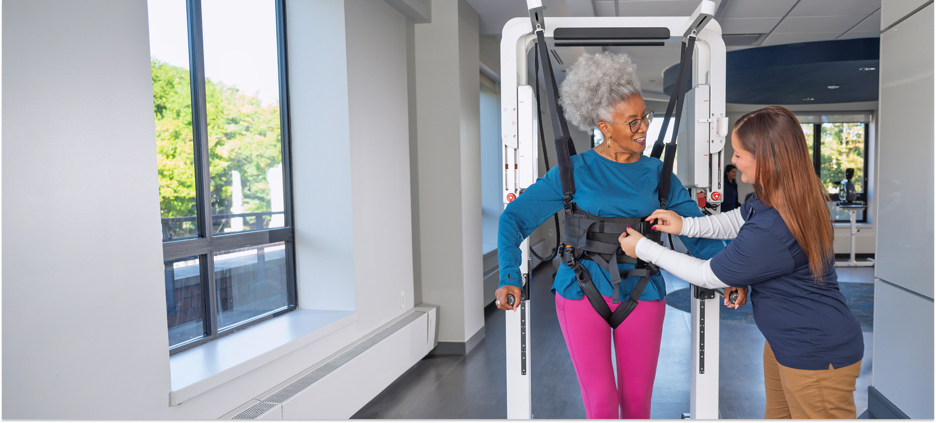 Older woman in walking harness assisted by physical therapist in a well-lit rehabilitation facility.