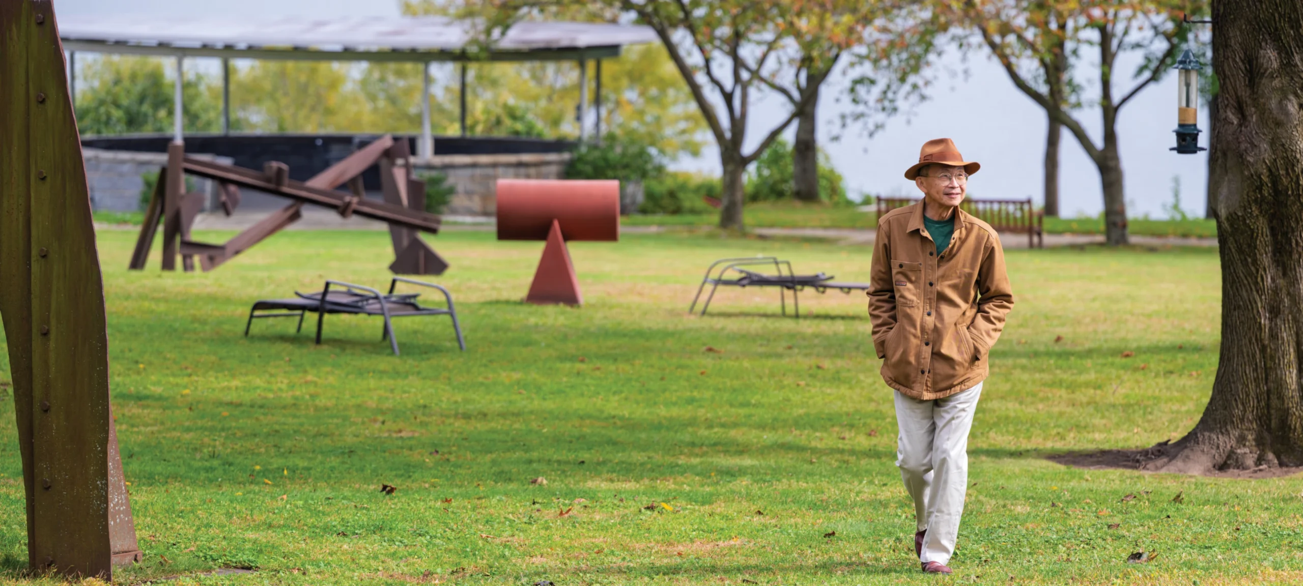 Man in brown jacket and hat walks through a park with sculptures, green grass, and surrounding trees.