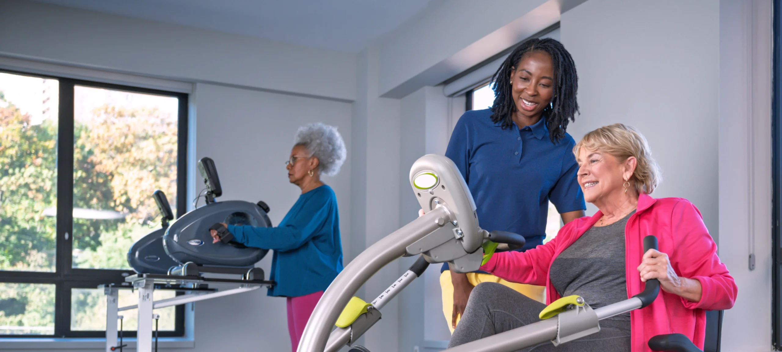 Trainer assists one of two women using exercise machines in a well-lit fitness room.