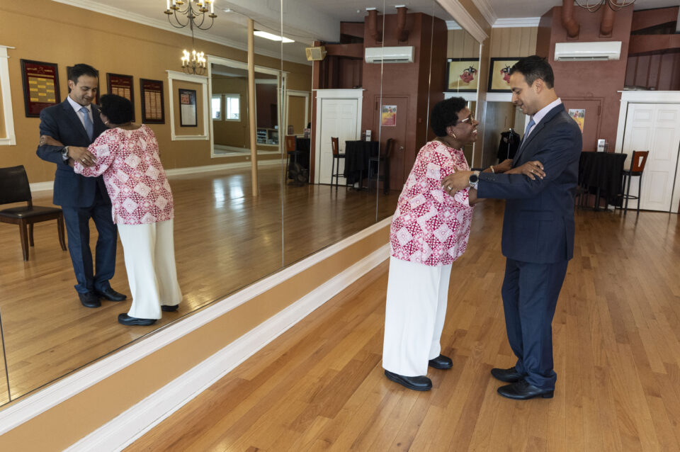 A man and woman stand facing each other, holding hands, and practicing dance steps on a golden dream dance floor in a mirrored studio with wooden floors.