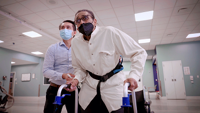 Man helps another man with a walker in a medical facility; both wear face masks.