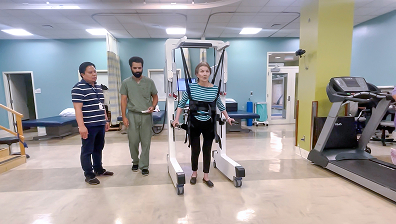Woman using a harness walker with support from two therapists in a rehabilitation center.