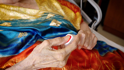 Elderly person in bed with a colorful blanket, pressing a call button with their right hand.