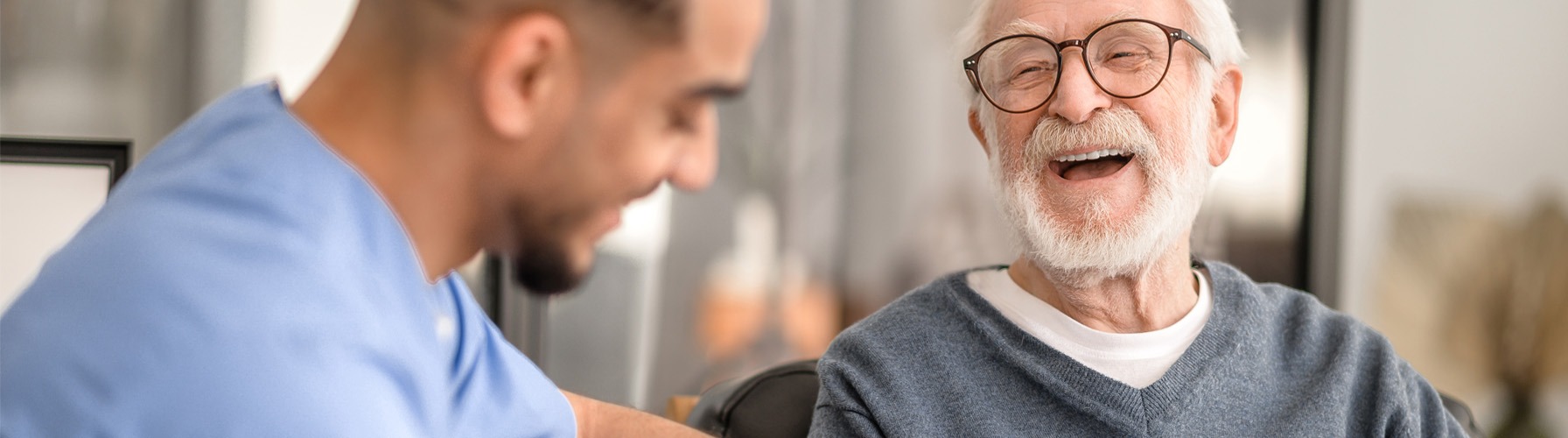 Smiling elderly man with glasses sits and talks to a caregiver in blue scrubs.