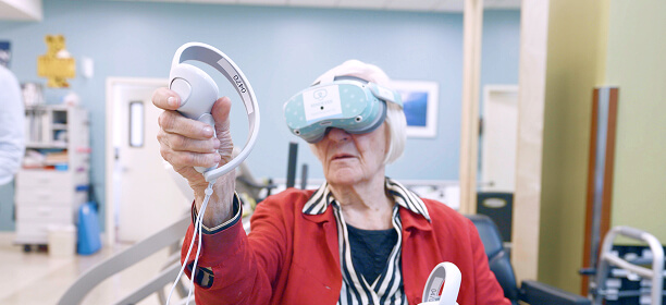 An older adult wearing a virtual reality headset and holding controllers in both hands participates in a short-term rehabilitation activity indoors.