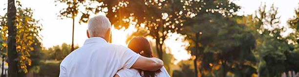 An older man and a younger woman walk outdoors at sunset, with the man’s arm around the woman’s shoulder near the Hebrew Home in Riverdale. Trees and sunlight are visible in the background.