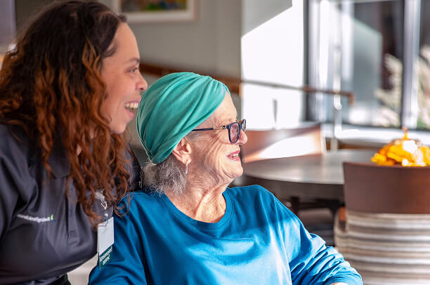 A younger woman and an older woman with a teal headscarf and glasses sit together, both smiling, at the sunlit Riverdale Hebrew Home, highlighting the warmth of senior care in this inviting indoor setting.