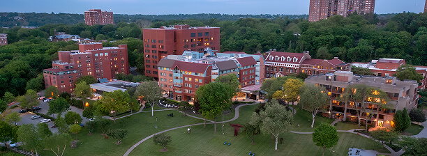 Aerial view of a campus with brick buildings, green lawns, trees, walking paths, and tall residential buildings in the background, featuring the Riverdale nursing home and facilities for senior care.