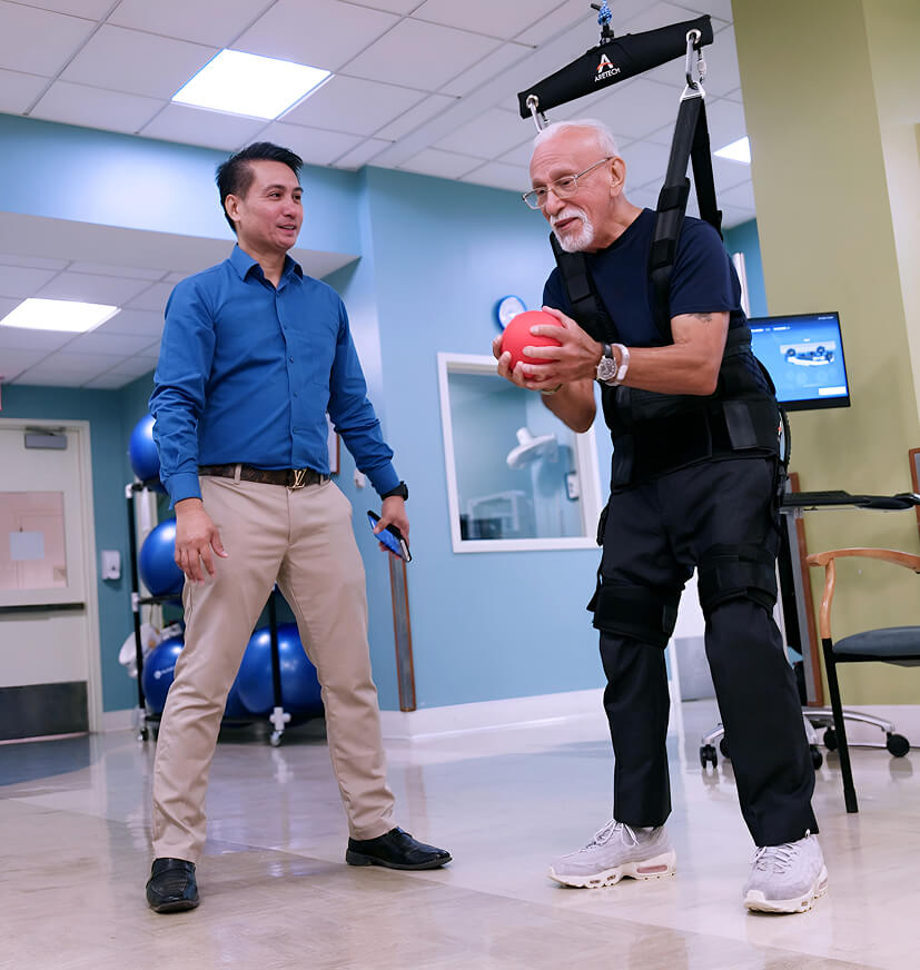 An older man in a physical therapy harness holds a red ball while a therapist in business casual attire observes at Hauser Rehabilitation Center.