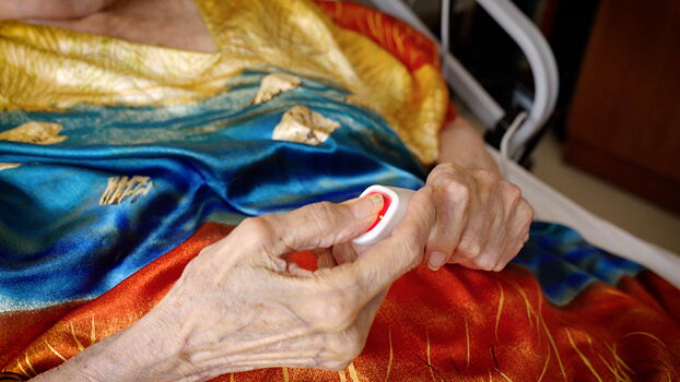 A resident at Riverdale's Hebrew Home in a colorful gown lies in bed, pressing a red emergency call button with one hand, highlighting attentive senior living care.