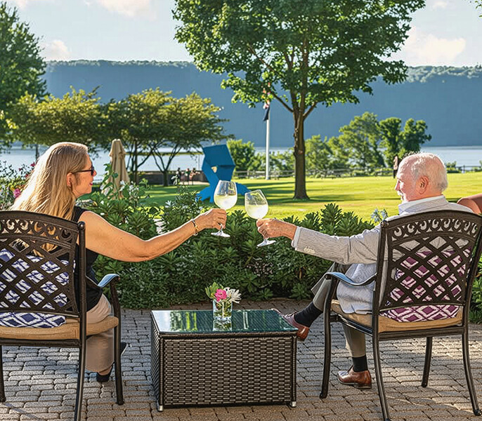 Two people sit on patio chairs clinking wine glasses at the Hebrew Home at Riverdale, enjoying a scenic landscape with trees, grass, and water in the background.