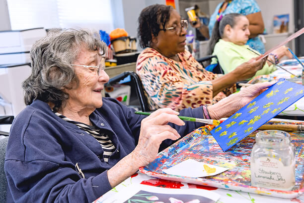 Three women sit at a table in a bright room at Hebrew Home in Riverdale, painting on canvases with brushes. Art supplies and a labeled jar are visible on the table, reflecting an engaging senior living activity.