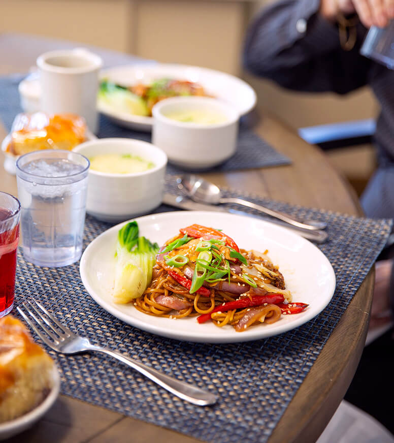 A plate of stir-fried noodles with vegetables and green onions is on a set dining table with soup, drinks, and bread at the Hebrew Home in Riverdale.