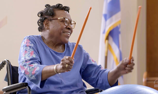 An older woman in a wheelchair smiles while holding two orange drumsticks, participating in a drumming activity at Hauser Rehabilitation Center. A flag is visible in the background.