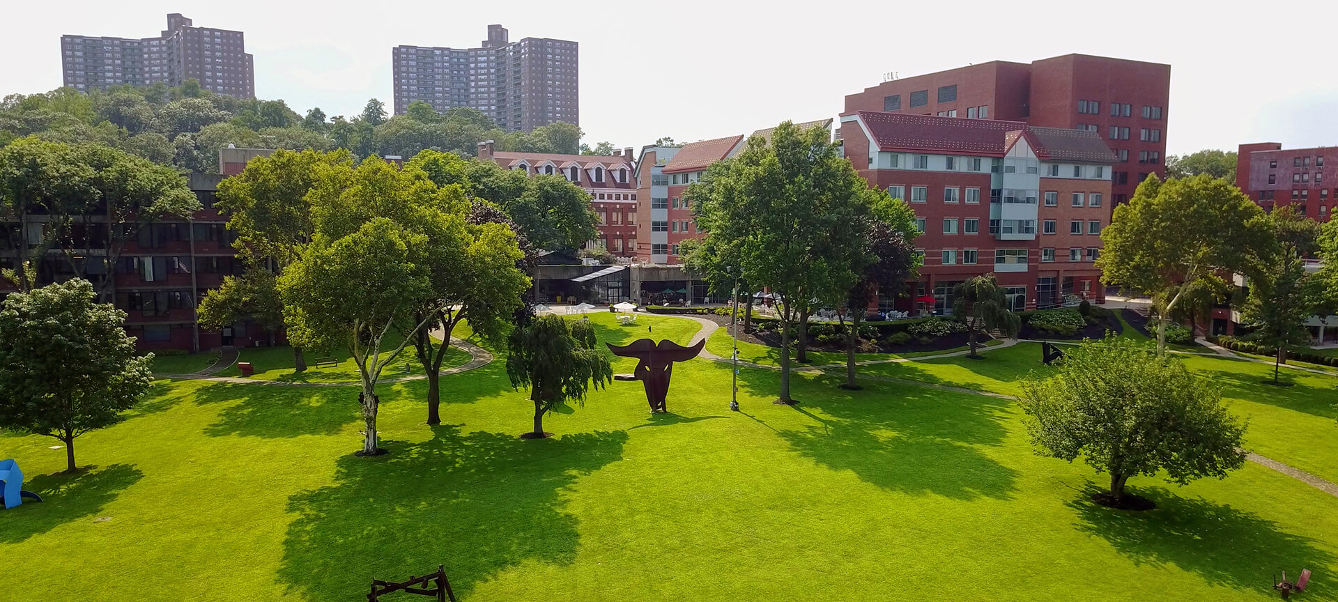 Green lawn with scattered trees, a large dark sculpture at the center, and red-brick buildings and tall apartments—including the Hebrew Home at Riverdale—in the background.