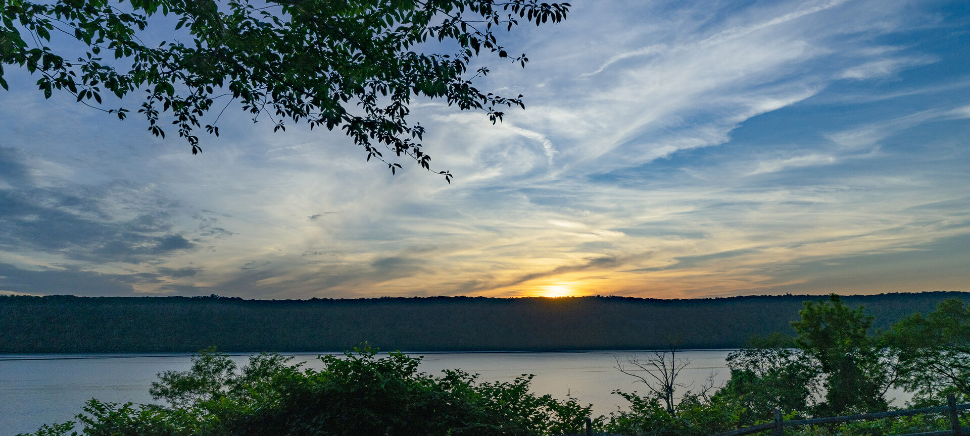 Sunset over a tranquil river with a tree-lined horizon, scattered clouds above, and autumn foliage in the foreground.