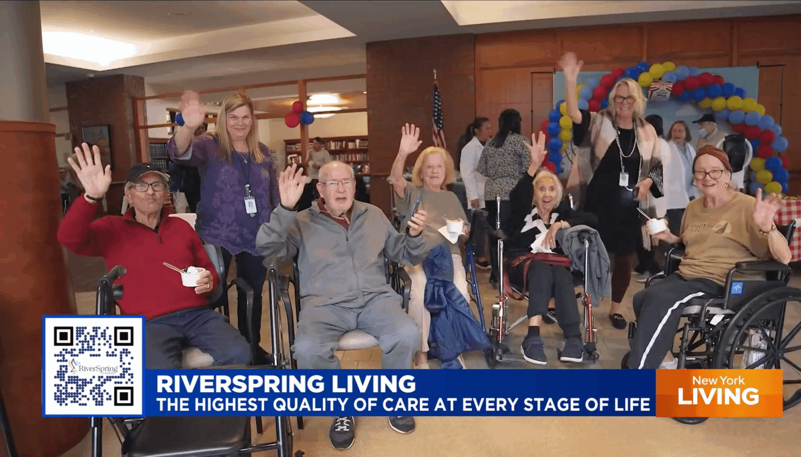 A group of older adults, caregivers, and community members smiling and waving at a Riverspring Living event, with balloons in the background and signs for Hudson Care and New York Living visible.
