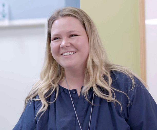A woman with long blonde hair wearing a navy blue blouse is smiling indoors, standing in front of a yellow and blue wall at a Short-Term Rehabilitation center.