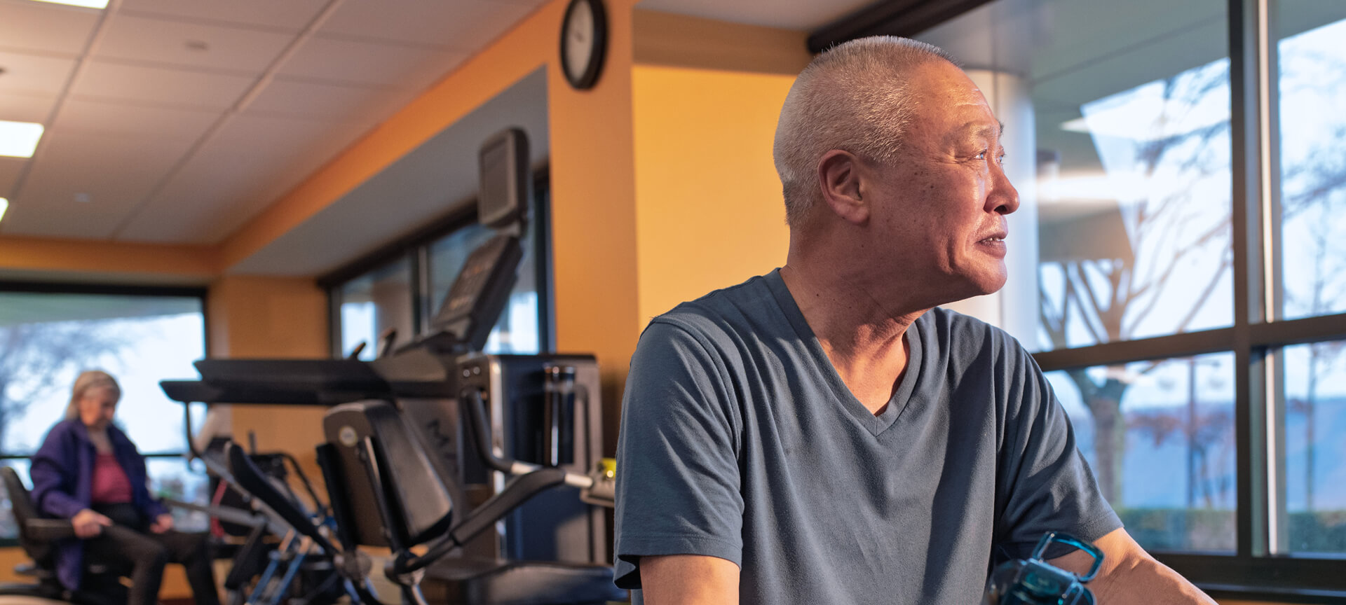 An older man sits in a gym near exercise equipment, looking to the side, while a woman is seated in the background. Large windows let in natural light, highlighting a welcoming space for Short-Term Rehabilitation.