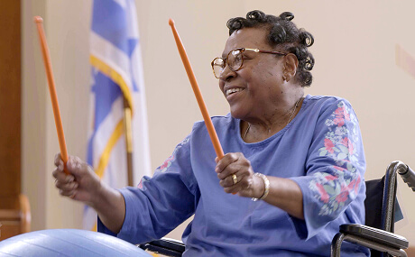 An older woman in a wheelchair smiles while holding two orange drumsticks, with a flag visible in the background—capturing the spirit of Short-Term Rehabilitation.