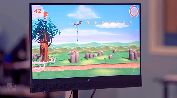 A computer screen displays a colorful side-scrolling game featuring a flying character collecting coins above a landscape with trees and rocks, offering engaging activities ideal for short-term rehabilitation.
