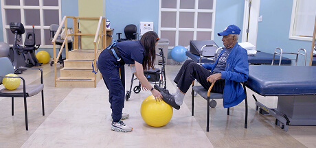 A physical therapist assists a man seated with a cane, helping him rest his foot on a yellow exercise ball in a short-term rehabilitation facility.