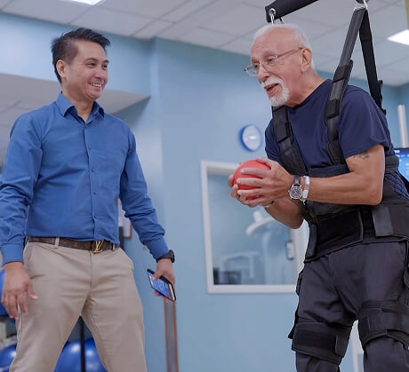An older man in a harness holds a red ball during Short-Term Rehabilitation, while a therapist in a blue shirt stands nearby and observes.