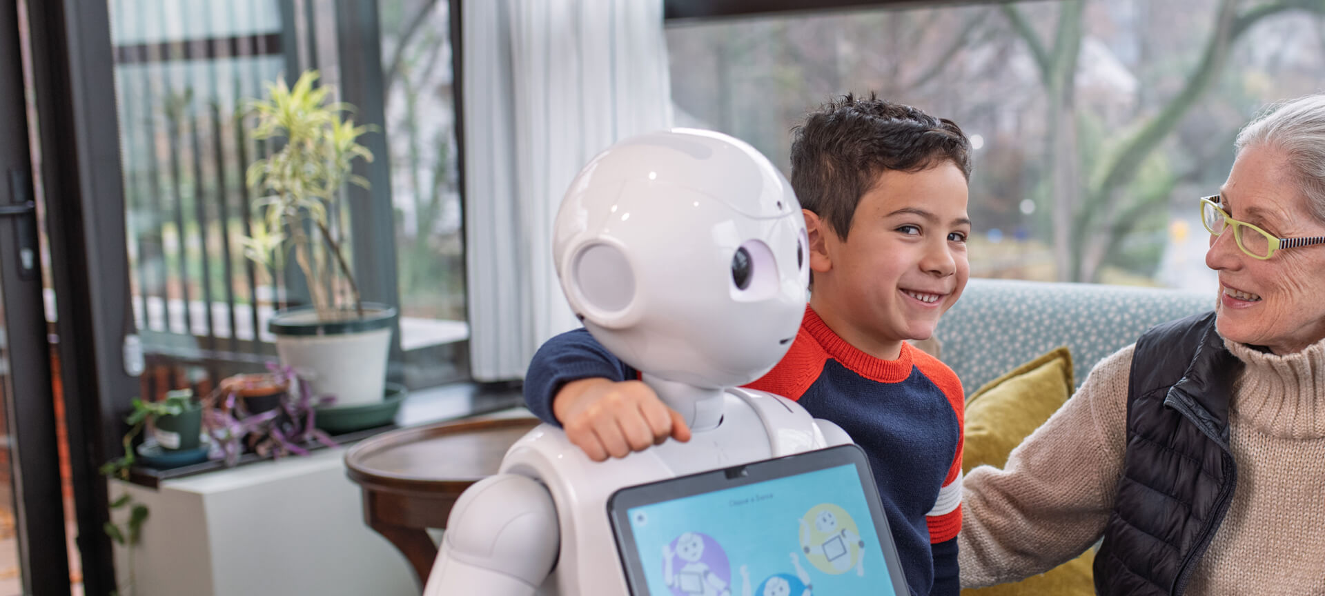 A boy with his arm around a humanoid robot sits next to an older woman in a living room filled with large windows and plants.