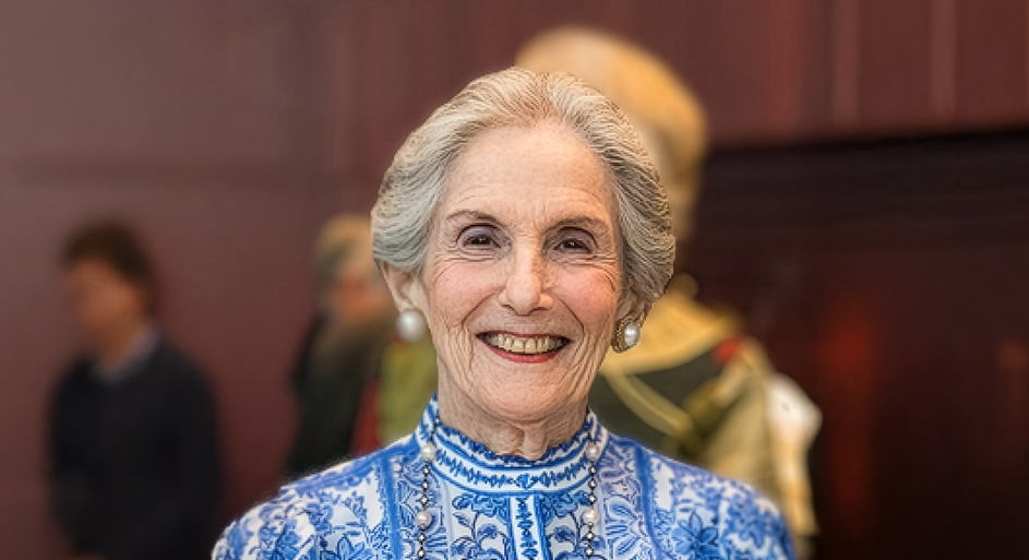 Gray-haired woman with pearl earrings smiles, wearing a blue and white patterned blouse, standing indoors with blurred people in the background.