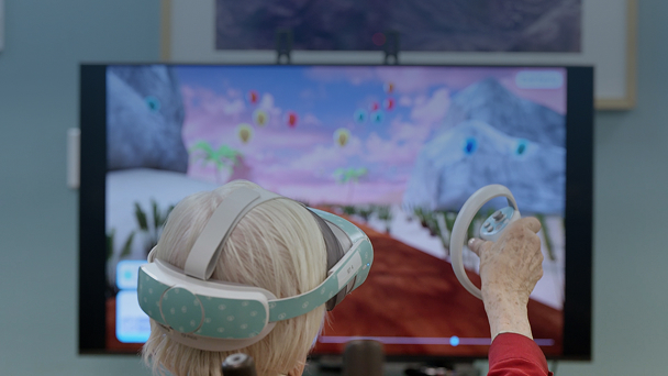An older adult wearing a virtual reality headset and holding a controller explores an immersive digital landscape displayed on a large screen.