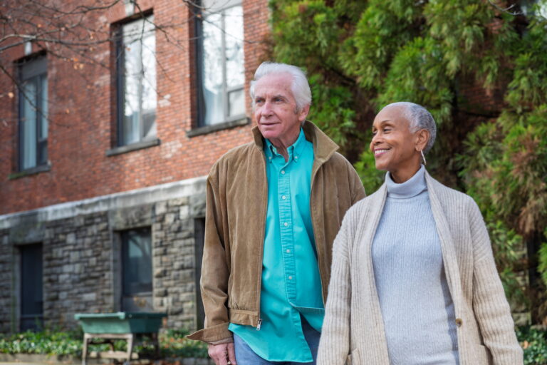 Older couple walks outdoors, smiling, in front of brick building and green trees.