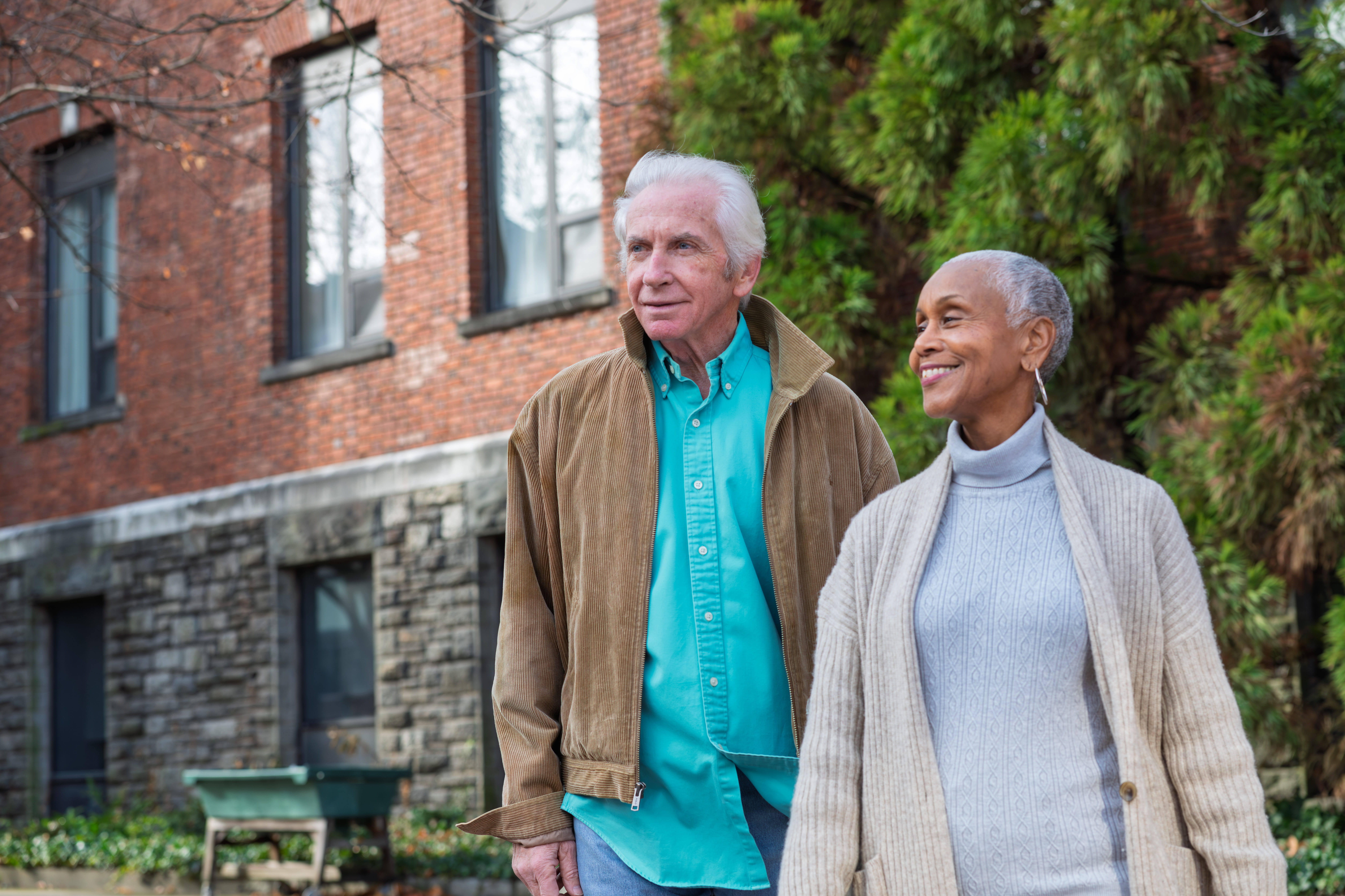 Older couple walks outdoors, smiling, in front of brick building and green trees.