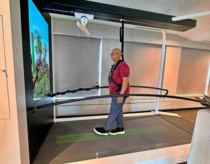 Older man walks on treadmill with safety harness, facing a virtual reality screen showing outdoor scenery.
