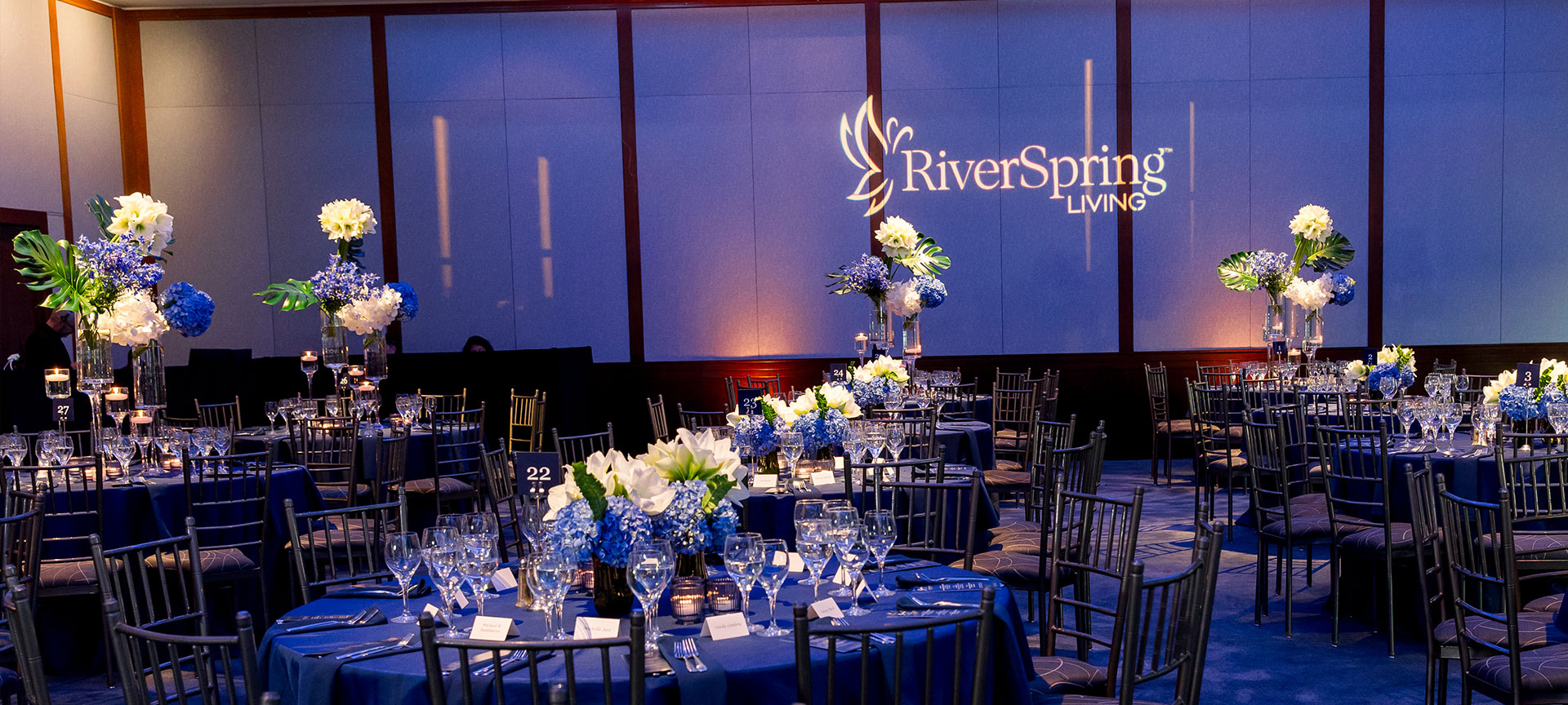 Event banquet hall with round tables in blue linens, floral centerpieces, glassware, and RiverSpring Living logo projected on wall.