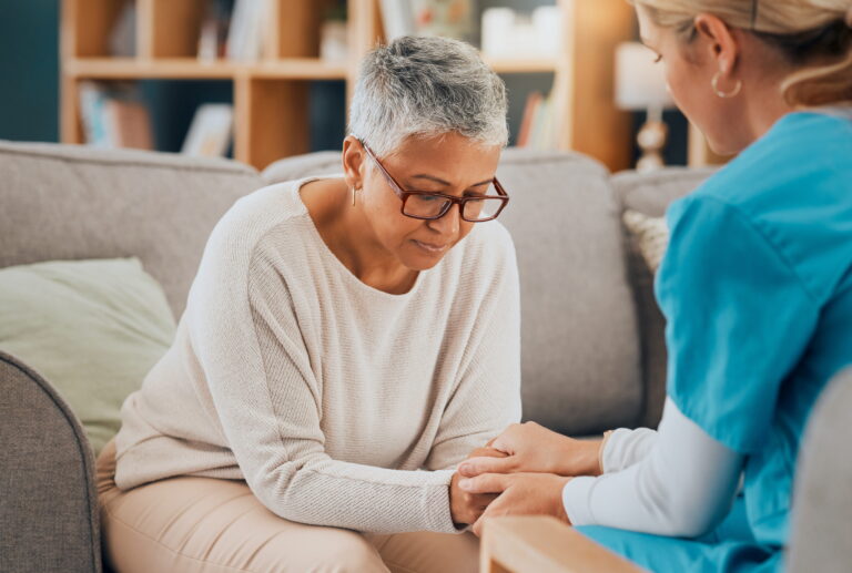 Older woman on couch holds hands with nurse in blue scrubs; both appear thoughtful and quiet.