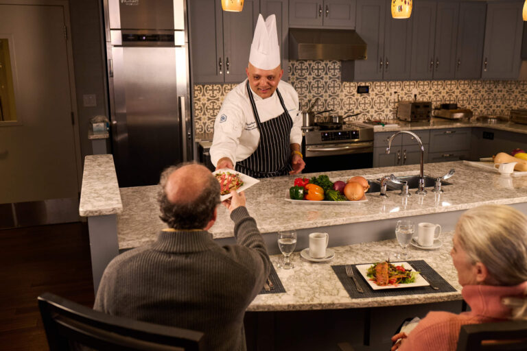 Chef in white hat serves meal to seated man in modern Bronx kitchen with vegetables on counter.