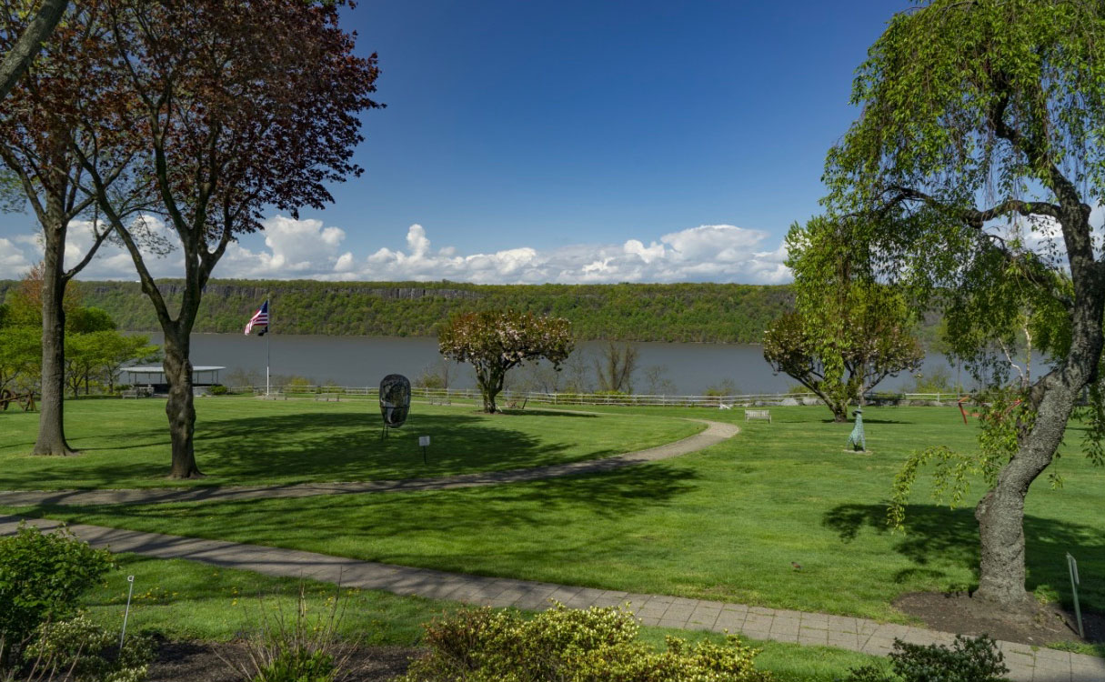 Riverside park in Riverdale with green grass, trees, a path, American flag, Hudson River, and hills.