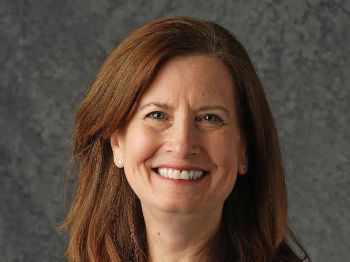 Woman smiling, shoulder-length brown hair, standing in front of a gray textured background.