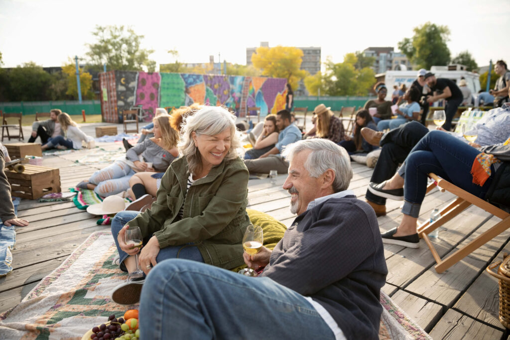 Two older adults sit on a blanket at an outdoor event in New York, smiling and holding drinks.