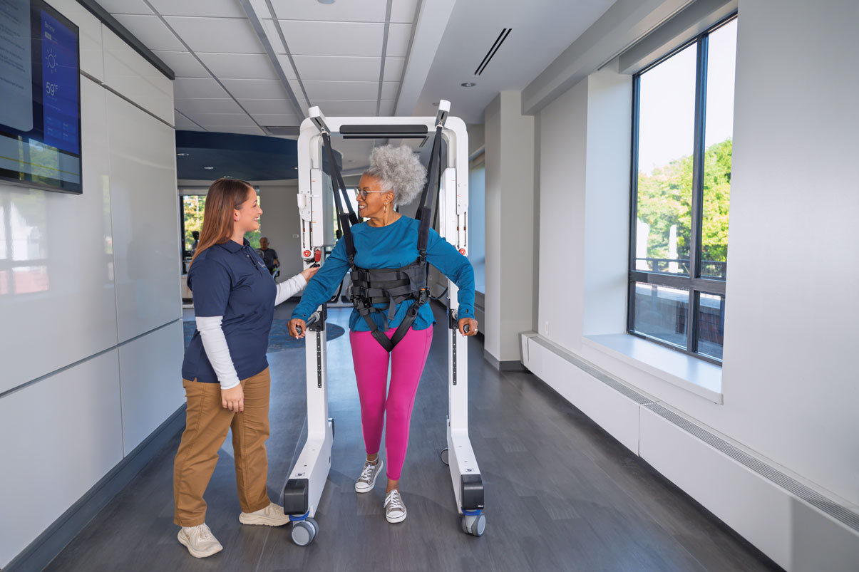 Older woman using robotic walker with healthcare worker assisting in a bright hallway.