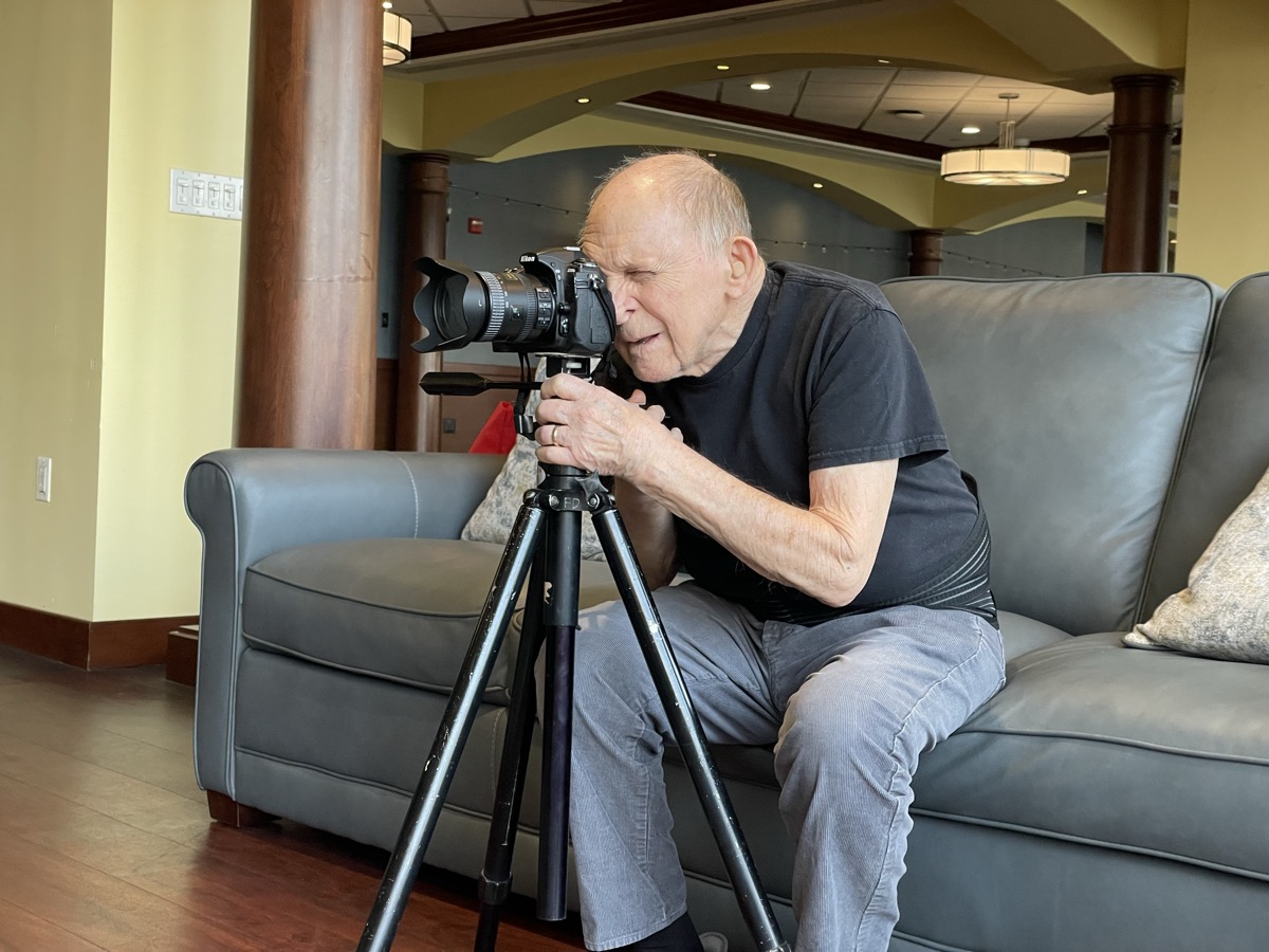 Elderly photographer sits on couch, using a tripod-mounted camera to take a photo.
