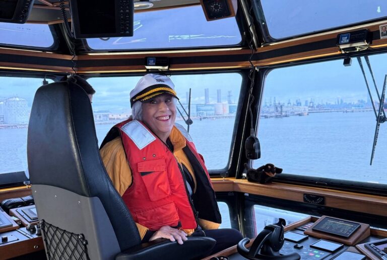 Retired teacher in captain's hat and life vest steering a tugboat, city and water visible outside.