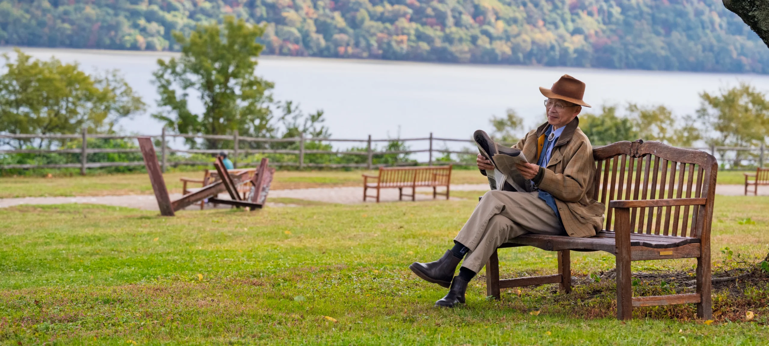 Elderly man in brown hat reads newspaper on lakeside bench; trees in the background.
