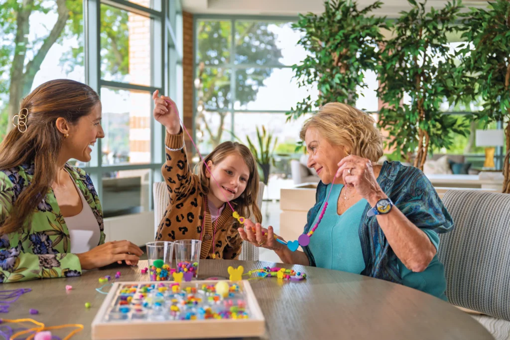 Three generations sitting at a table, smiling as they make colorful bead jewelry together.