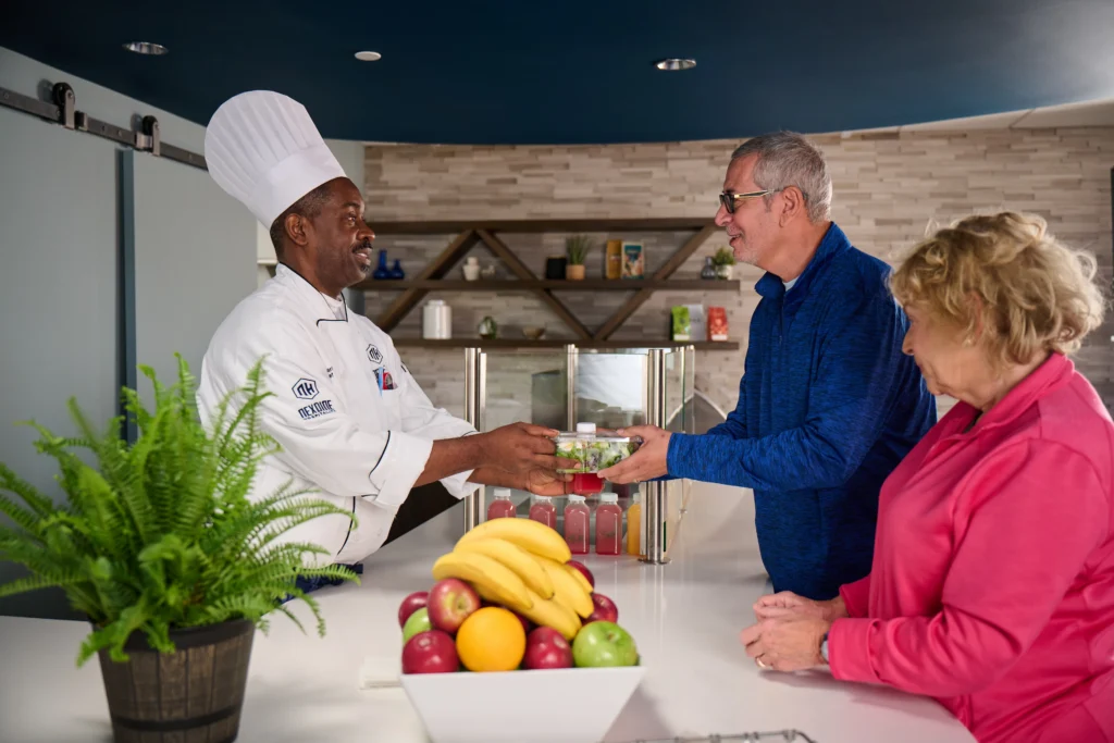 Chef gives salad to man as woman stands beside them at a counter with fruit and a fern.
