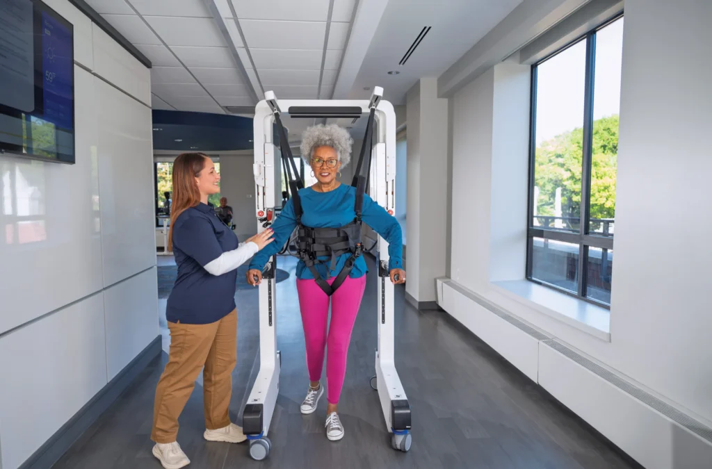 Older woman in walking harness for rehabilitation, assisted by healthcare worker in a bright, well-lit room.
